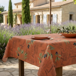 Könnte beinhalten: Eine terrakottafarbene Tischdecke mit einem Provence-Design mit Olivenzweigen und Oliven. Die Tischdecke liegt auf einem Holztisch mit einem Teller, einem Löffel und Weingläsern. Eine Schüssel mit Oliven ist ebenfalls vorhanden.