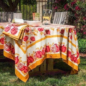 Könnte beinhalten: Ein runder Tisch für ein Abendessen im Freien, bedeckt mit einer weißen Tischdecke mit einem sich wiederholenden Muster aus roten Granatäpfeln und gelben Rändern. Servietten und eine Schüssel stehen auf dem Tisch, mit einem Krug und Gläsern.