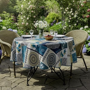 Könnte beinhalten: Ein runder Esstisch im Freien mit einer gemusterten Tischdecke in Blau, Weiß und Gold. Der Tisch ist mit Tellern, Gläsern und einer Vase mit Blumen gedeckt. Zwei Korbstühle sind um den Tisch herum aufgestellt.