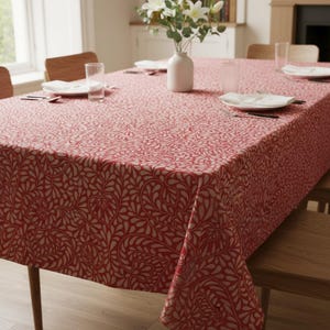 Könnte beinhalten: Eine rechteckige rote Tischdecke mit weißem Blumenmuster bedeckt einen Holztisch. Der Tisch ist mit weißen Tellern, Besteck und klaren Gläsern gedeckt. Eine weiße Vase enthält weiße Blumen.