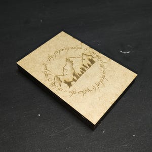 May include: A rectangular card with a tan, textured surface featuring a dark brown laser-engraved design. The artwork depicts a silhouette of a group of figures walking through a mountain pass, encircled by a ring of intricate, flowing script.