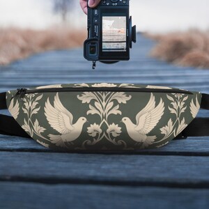 May include: A dark green fanny pack with a cream-colored dove and floral pattern. The bag has a black adjustable strap and a black zipper. The fanny pack is photographed outdoors on a wooden walkway.