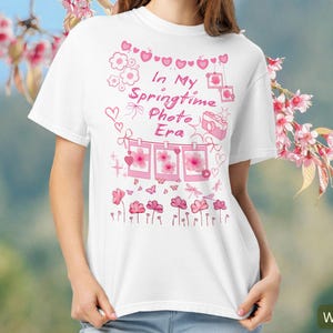 Könnte beinhalten: Weißes T-Shirt mit rosa Grafikdesign. Das Design enthält den Text "In My Springtime Photo Era" mit rosa Blumen, Herzen und einer Kamera-Illustration. Das Shirt ist ein lässiger Stil mit kurzen Ärmeln.