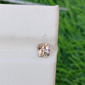 May include: A square-cut, peach-colored gemstone with a faceted surface. The gem is set against a white, textured background, with a blurred green backdrop. The gemstone's color ranges from light peach to a darker, more saturated tone.