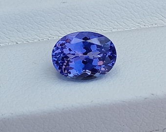 May include: Oval-shaped, faceted tanzanite gemstone. The gem displays a deep blue-violet color with internal reflections. It is set against a white background, showcasing its clarity and brilliance. A beautiful example of a precious stone.