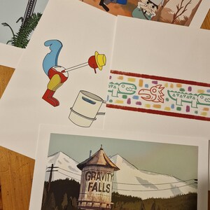May include: Four different prints featuring characters and imagery from the animated television series Gravity Falls. The prints include a stop sign, a character with a frog on their head, a bird with a bucket, and a water tower with the words "Gravity Falls" on it.