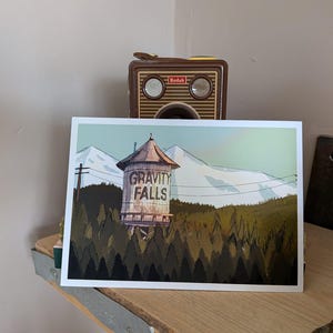 May include: A print depicting the Gravity Falls water tower, set amidst a forest and mountains. The illustration includes the text "GRAVITY FALLS" on the water tower. The print is displayed on a wooden shelf.