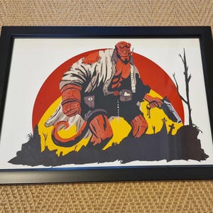 May include: Framed art print featuring a red-skinned demon-like character with a gun, set against a red sun and yellow landscape. The artwork is in a comic book style, with a black frame. The image includes a graveyard scene.