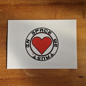 May include: A white card with a black and red circle design. The circle says "In Space We Trust" with a red heart in the center.