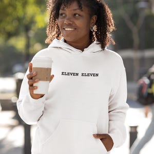 Op de afbeelding: Een witte hoodie met de tekst "ELEVEN ELEVEN" in het zwart. De hoodie heeft een voorzak en een capuchon met trekkoord. De persoon op de foto houdt een witte koffiebeker met een bruine hoes vast.