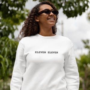 Op de afbeelding: Witte crewneck sweatshirt met de tekst "ELEVEN ELEVEN" in het zwart op de voorkant.