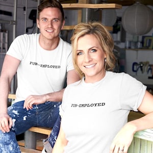 May include: Two people wearing white t-shirts with the text "FUN-EMPLOYED" printed on the front. The man is wearing blue jeans and the woman is wearing a white t-shirt.