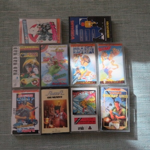 May include: A collection of 10 vintage video game cartridges for the Amstrad CPC computer. The cartridges are in clear plastic cases with colorful artwork featuring characters and scenes from the games. The titles include "Mad Mad Mad", "Los Angeles SWAT", "Skyfox", "Saigon Combat Unit", "Bump", "Milk Race", "Vent D'mar", "Your Head", "Phaser", and "Wordhang".