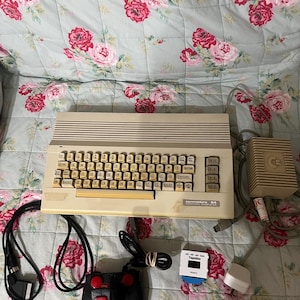 Peut inclure: Un ordinateur personnel Commodore 64 vintage, avec un clavier beige et un joystick noir avec des boutons rouges. L'ordinateur est accompagné d'une alimentation et d'autres accessoires. Le clavier a des touches jaunes.
