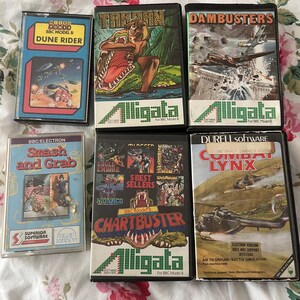 以下が含まれることがあります： ヴィンテージのBBC Model Bコンピューターゲームカートリッジのコレクション。「Dune Rider」、「Tarzan」、「Dambusters」、「Smash and Grab」、「Chartbuster」、「Combat Lynx」などのタイトル。ゲームボックスにはカラフルなグラフィックとテキストが表示され、「Alligata」のロゴが付いています。