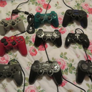 Op de afbeelding: Een verzameling van negen verschillende videogamecontrollers, waaronder zwarte, grijze, rode, blauwe en groene controllers. Sommige controllers hebben logo's en tekst erop, zoals "E-MU-S" en "Logitech".