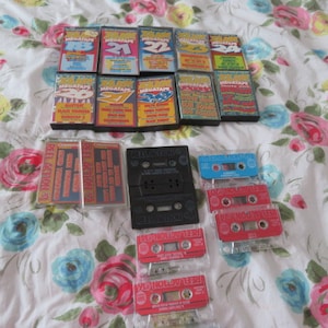 Può includere: Una collezione di cassette audio con vari titoli, tra cui "Megatape" e "Action", esposte su un tessuto bianco con motivo floreale. Le cassette sono di vari colori, tra cui rosso, nero e bianco.