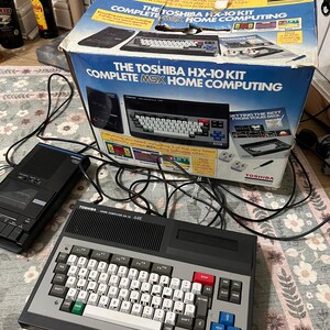 Puede incluir: Un kit de ordenador doméstico MSX Toshiba HX-10 vintage, con teclado, reproductor de casetes y caja original. La caja dice "The Toshiba HX-10 Kit Complete MSX Home Computing". El teclado es gris y negro con teclas blancas.