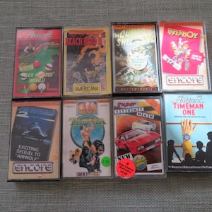 May include: Eight vintage cassette tapes for a video game console. The tapes are in various colors and feature different game titles, including "Alex Higgins' World", "Beach Head", "The Chuckle Brothers Show", "Paperboy", "Airwolf 2", "Zzap! Test Drive", "Super Stuntman", and "Timeman One".