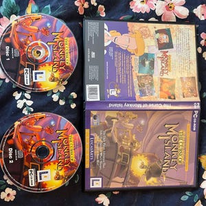 Puede incluir: Dos cajas de CD para el videojuego The Curse of Monkey Island. La parte delantera de la caja presenta a un pirata de dibujos animados sosteniendo una espada y un mono. La parte posterior de la caja tiene una descripción del juego y una lista de características.