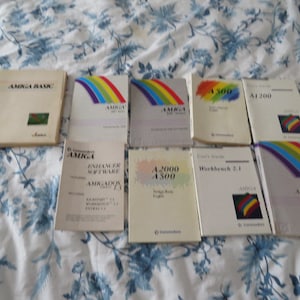 May include: A collection of ten vintage Amiga computer manuals, including titles like "Amiga Basic", "Workbench 2.1", "Using the Amiga Workbench", and "A2000 A500". The manuals are in various colors, including white, blue, and green, and feature rainbow stripes.