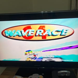 May include: A TV screen showing "Wave Race 64" in red and blue. A Nintendo 64 console, a grey controller, and two game cartridges are on a wooden surface below the screen. The console is black.