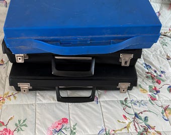 May include: A stack of two briefcases, one black and one blue. The blue briefcase is on top, with a handle and a latch. The black briefcase is underneath, with a handle and a silver latch. The briefcases are on a floral patterned bedspread.