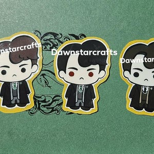 May include: Three die-cut stickers featuring chibi-style characters with dark hair, wearing black coats over grey vests and white shirts with green ties. Each sticker has a yellow border and the text Dawnstarcrafts printed across the center.