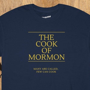 Op de afbeelding: Een donkerblauw t-shirt met de tekst "THE COOK OF MORMON" in gouden letters. De tekst "MANY ARE CALLED, FEW CAN COOK" staat ook in gouden letters onder de hoofdtekst.