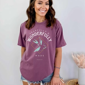 Woman wearing a Berry Comfort Colors T-shirt with a teal sparrow in an orange diamond surrounded by the text "FEARFULLY & WONDERFULLY MADE PSALM 139"