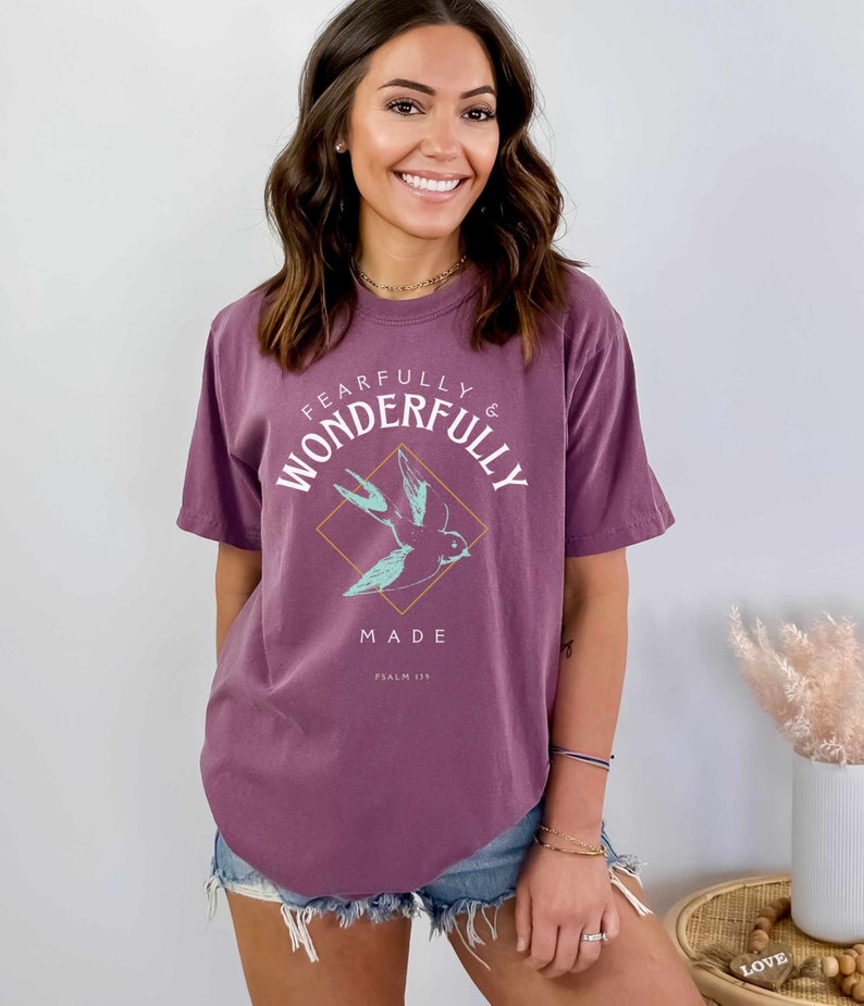Woman wearing a Berry Comfort Colors T-shirt with a teal sparrow in an orange diamond surrounded by the text "FEARFULLY & WONDERFULLY MADE PSALM 139"
