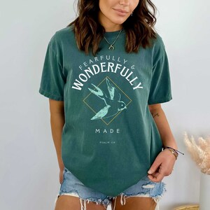 May include: Teal blue t-shirt with a white graphic of a bird in flight inside a diamond shape. The text "Fearfully & Wonderfully Made" is written around the diamond and "Psalm 139" is written below the diamond.