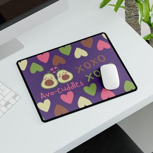 May include: Purple mousepad with a black border featuring a design of two avocado halves with hearts and the text "Avo-cuddles" and "XOXO". A white mouse and keyboard are on a white desk, with a computer monitor displaying the time.
