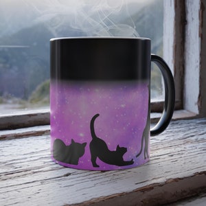 Puede incluir: Una taza negra que cambia de color con siluetas de gatos sobre un fondo de galaxia púrpura. La taza es negra cuando está fría y revela el diseño de los gatos cuando está caliente. Sale vapor de la taza, lo que sugiere una bebida caliente.