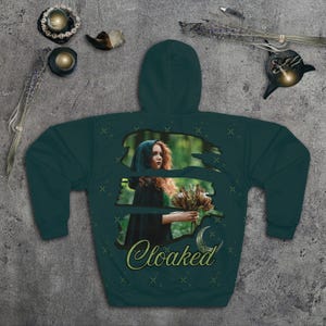May include: A dark teal hoodie featuring a graphic of a person in a cloak holding flowers, with the word "Cloaked" in a script font. The design is set against a forest backdrop with a crescent moon and stars.