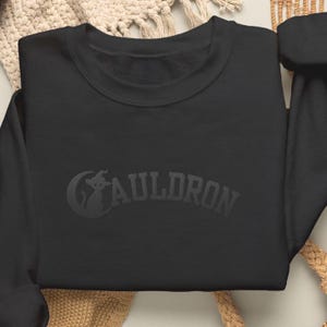 May include: A black long-sleeve crewneck sweatshirt featuring a tonal black embroidered design on the chest. The graphic includes a silhouette of a cat wearing a witch hat positioned inside a crescent moon next to the word CAULDRON in block letters.