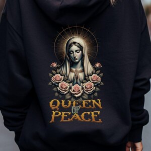 Könnte beinhalten: Schwarzer Kapuzenpullover mit einem Aufdruck einer Frau mit Heiligenschein, Rosen und dem Text "QUEEN OF PEACE" in Gold. Der Hoodie hat eine Kapuze mit Kordelzug und lange Ärmel. Das Bild befindet sich auf der Rückseite.