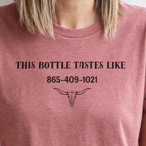 May include: A maroon t-shirt with the text "THIS BOTTLE TASTES LIKE 865-409-1021" and a stylized bull skull graphic.