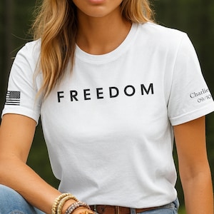 May include: White t-shirt with the word "FREEDOM" printed in black across the chest. The American flag is printed on the left sleeve, and the name "Charlie" with the date "09/10" is printed on the right sleeve. The person is wearing blue jeans and bracelets.