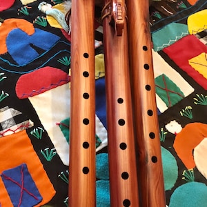 May include: Three wooden flutes with carved animal figures and multiple finger holes. The flutes are a warm brown color and rest on a colorful patchwork quilt. A green cloth is in the background.