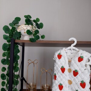 May include: White crocheted vest with a strawberry pattern and wooden buttons. The vest is hanging on a hanger on a wooden shelf with two gold candle holders.