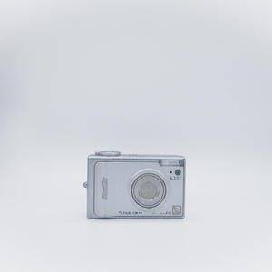 May include: A silver compact digital camera featuring a central lens, a small flash unit in the upper right corner, and the text FujiFilm printed on the front body. The device has a rectangular shape with rounded edges and a shutter button on top.