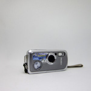 Op de afbeelding: Een zilveren Pentax digitale camera met een 5x optische zoom en waterdichte functies. De camera heeft een zwarte band.