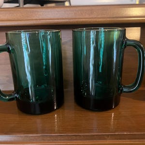 Può includere: Due tazze di vetro verde smeraldo con grandi manici. Le tazze hanno una forma rettangolare e una base verde scuro. Le tazze sono esposte su una superficie di legno.
