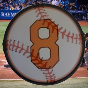 May include: A circular baseball-themed sign featuring the number 8 in orange, set against a white background with red stitching details. The sign has a black border and is likely a sports-related item.