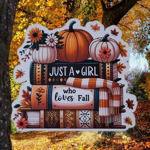 May include: A die-cut sticker featuring a stack of three books topped with two pumpkins and a vase of flowers. The top book reads "JUST A GIRL" and the middle book reads "who loves Fall". A striped scarf is draped over the books, accented by autumn leaves.