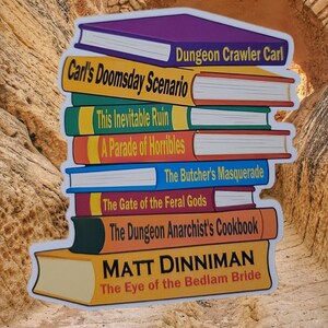 Puede incluir: Una pila de pegatinas de novelas de fantasía con títulos como "Dungeon Crawler Carl" y "Carl's Doomsday Scenario". Los libros tienen lomos coloridos y páginas blancas. También es visible el nombre del autor "MATT DINNIMAN".