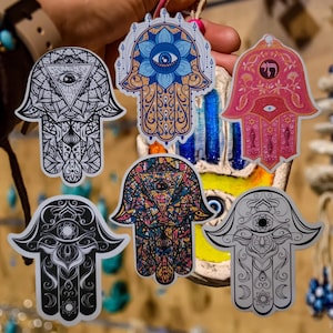 Könnte beinhalten: Sechs bunte Hamsa-Hand-Aufkleber mit komplizierten Designs und einem blauen bösen Auge in der Mitte. Die Aufkleber befinden sich auf einem weißen Hintergrund.