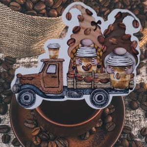May include: A die-cut sticker featuring a brown vintage truck carrying two gnomes with coffee-themed hats and cups. One gnome holds a cup labeled COFFEE. The design is decorated with scattered coffee beans and features a warm brown color palette.