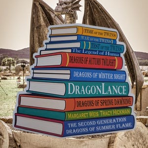 以下が含まれることがあります： 「Dragonlance」や「Dragons of Autumn Twilight」などのタイトルが書かれたカラフルな本の切り抜きが積み重ねられています。本はドラゴンの像の前にあります。タイトルは本の背表紙に白い文字で書かれています。